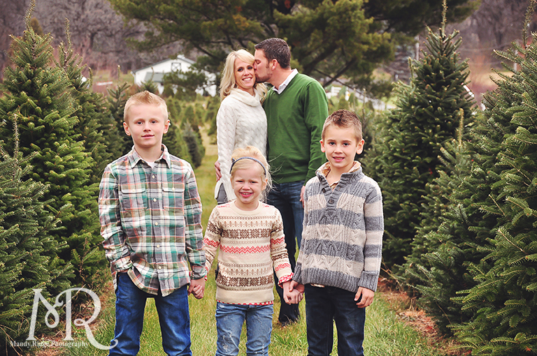 Family Christmas Portrait // Christmas Tree Farm // standing in between rows of trees with the kids in front and the parents in back kissing // by Mandy Ringe Photography