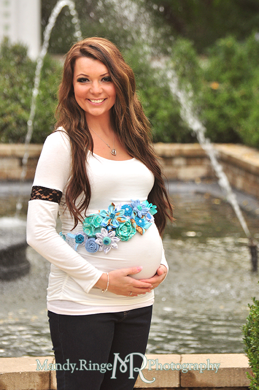 Pregnant woman posing in front of a fountain, holding her belly and wearing a handmade blue maternity sash // Maternity portraits // Hurley Gardens - Wheaton, IL // by Mandy Ringe Photography